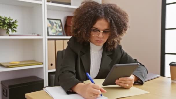 Зосереджена іспаномовна жінка з кучерявим волоссям робить нотатки в офісі - Кадри, відео