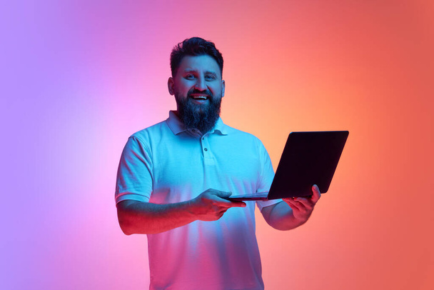 Knappe jongeman glimlachend kijkend naar de camera en houdt laptop in neon licht tegen levendige gradiënt studio achtergrond. Concept van menselijke emoties, zelfexpressie, werken en studeren online. Steun - Foto, afbeelding