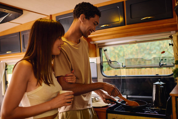 Ein gemischtrassiges Paar bereitet freudig eine gemeinsame Mahlzeit in einem gemütlichen Wohnmobil zu. - Foto, Bild