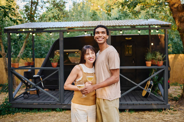 Una pareja interracial sosteniendo copas de vino, disfrutando de un momento romántico en su caravana durante una escapada. - Foto, Imagen