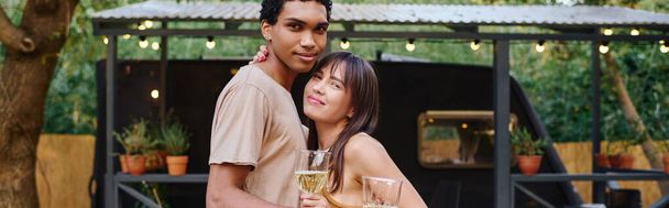 Ένας άντρας και μια γυναίκα, ένα διαφυλετικό ζευγάρι, κρατώντας ποτήρια κρασί, μοιράζονται μια ρομαντική στιγμή σε ένα τροχόσπιτο το ηλιοβασίλεμα.. - Φωτογραφία, εικόνα