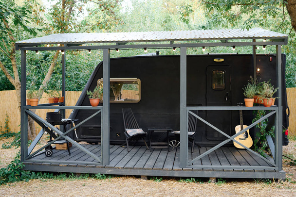 Um pequeno trailer preto com uma guitarra no alpendre, situado em um cenário sereno, evoca um momento musical acolhedor - Foto, Imagem