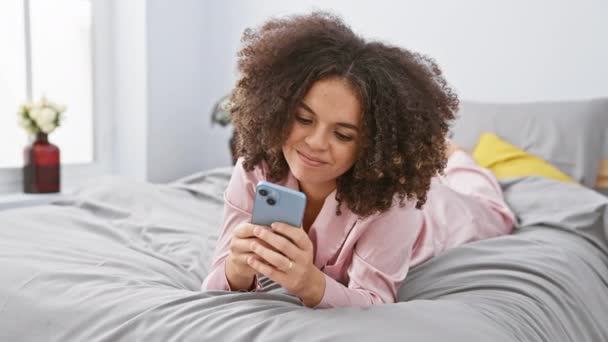 Fröhliche hispanische Frau mit lockigem Haar liegt im Bett, Smartphone in der Hand, gestikuliert hervorragend ok Zeichen, blinkt ein freundliches Lächeln umrahmt von der gemütlichen Schlafzimmer Hintergrund - Filmmaterial, Video