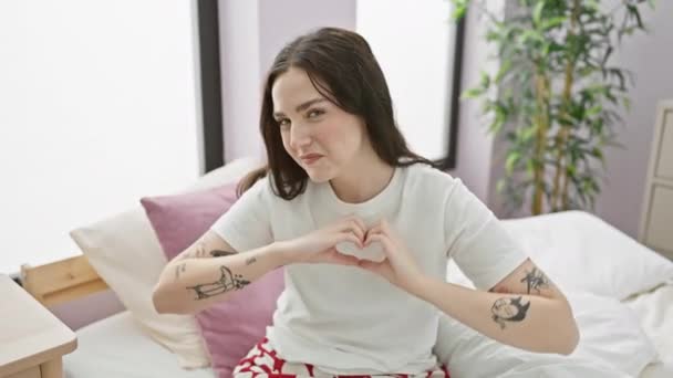 Cheerful young woman in pyjamas sits on bed, forming a heart symbol with her hands in a romantic gesture of love. captured in the cozy comfort of her bedroom. - Footage, Video