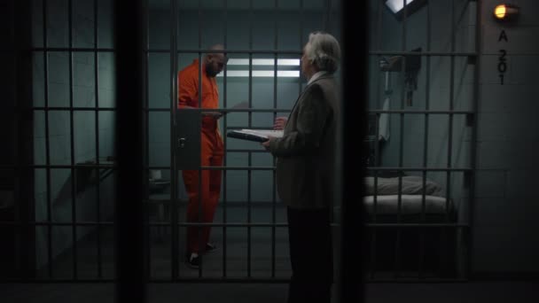 Więzień w pomarańczowym mundurze chodzi za kratkami w celi, rozmawia z adwokatem, czyta umowę adwokacką. Przestępca odsiaduje karę więzienia za przestępstwo w więzieniu. Centrum zatrzymań lub zakład karny. - Materiał filmowy, wideo