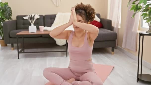Een jonge vrouw met krullend haar die yoga beoefent in een gezellige woonkamer, die rust en welzijn uitbeeldt. - Video