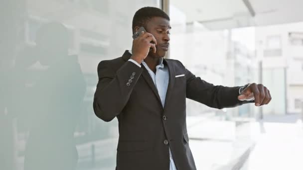 Уверенный, хорошо одетый африканский мужчина проверяет свои наручные часы, делая телефонный звонок в современной городской среде. - Кадры, видео