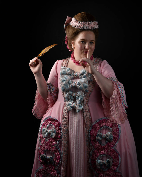 portrait of female model wearing an opulent pink gown,  costume of a historical French baroque nobility, style of Marie Antoinette. Holding feather quill. isolated silhouette on isolated dark studio background - Photo, Image