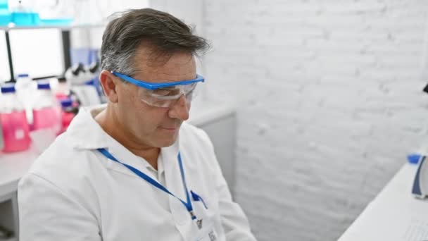 Een volwassen mannelijke wetenschapper met veiligheidsbril en labjas zit contemplatief in een modern chemisch laboratorium. - Video