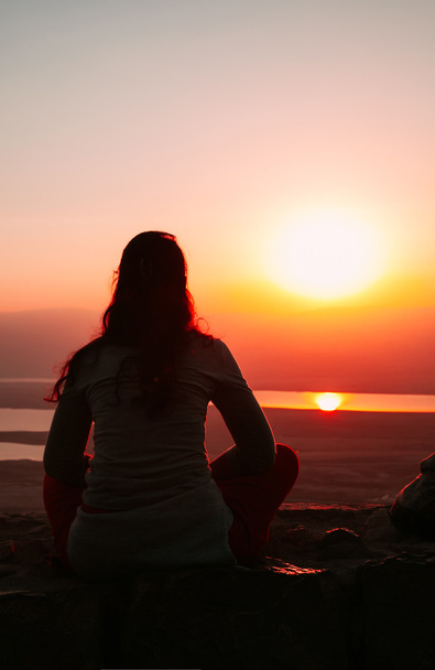 Silhouette einer Frau, die auf dem Boden sitzt und in den Sonnenuntergang blickt - Foto, Bild
