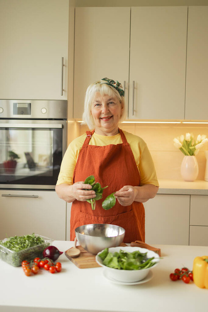 Senior 60 anos loira caucasiana sorrindo mulher segurando uma faca e espinafre fresco inteiro preparar uma tigela de salada de dieta saudável em sua cozinha rústica eco. Foto de alta qualidade - Foto, Imagem