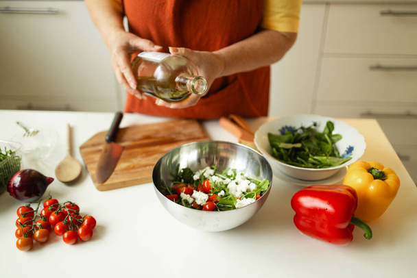 女性はキッチンで食べ物を準備しながら野菜サラダを季節ごと食べます. 認識できないシェフは自宅でベジタリアン料理を準備しながらオリーブオイルを加えます. 健康的な食事のコンセプト.  - 写真・画像