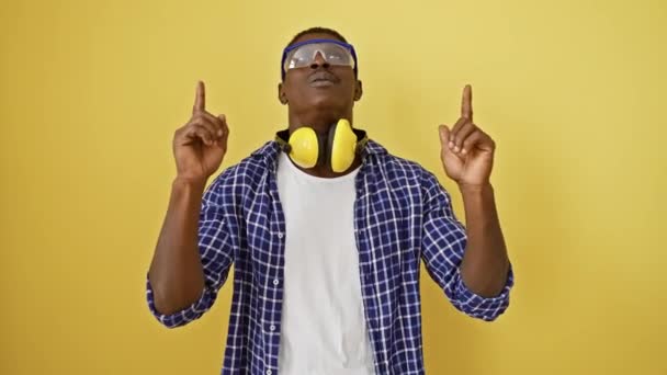 Vreugdevolle Afro-Amerikaanse man met veiligheidsbril kijkt verbaasd op, wijzend en verbaasd. alleen staan op een felgele geïsoleerde achtergrond, positieve vibraties uitstralend. - Video