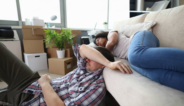 Porträt eines erschöpften Ehepaares, das es sich auf einem gemütlichen Sofa in einer neu erworbenen Wohnung gemütlich macht. Gestresstes Paar fühlt sich nach dem Umzug müde. Kartons mit Sachen auspacken. Umsiedlungskonzept - Foto, Bild