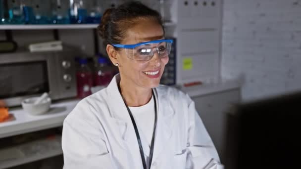 Mujer científica hispana burbujeante de mediana edad con gafas de seguridad apuntando con entusiasmo y sonriendo a la pantalla de la computadora en el laboratorio - Metraje, vídeo