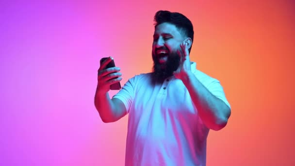 Чоловік в повсякденному вбранні тримає телефон і показує вітальний жест в неоновому світлі проти градієнтного рожево-оранжевого студійного фону. Концепція людських емоцій, соціальних медіа, мережі, зв'язок на відстані. - Кадри, відео