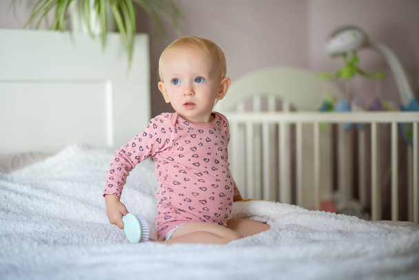 Toddler sitting on a bed with a hairbrush, crib in the background - Photo, Image