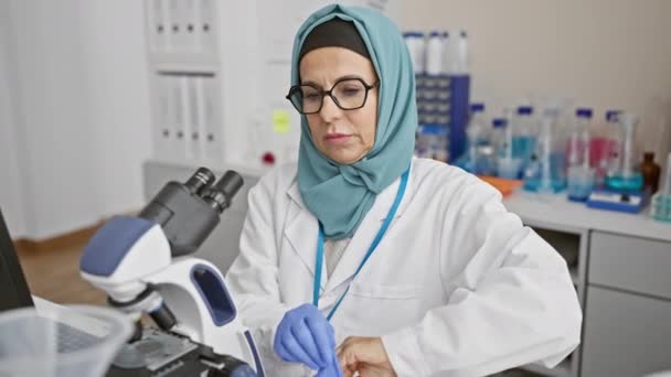Жінка середнього віку донорські рукавички на лабораторній лавці з мікроскопом та хімікатами, передаючи професіоналізм в охороні здоров'я. - Кадри, відео
