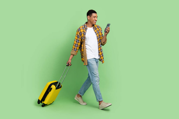 Full size profile photo of handsome guy walk hold suitcase use smart phone isolated on green color background. - Fotoğraf, Görsel