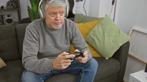 Starší muž s šedivými vlasy hraje videohry a reaguje emocionálně v útulném obývacím pokoji, ukazuje volný čas a zábavu doma. - Záběry, video