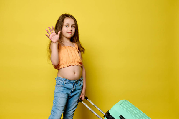 Adorable little traveler girl carrying blue suitcase, waving hello with her hand, looking at camera, isolated over yellow studio background. Copy advertising space. Childhood. Travel. Summer holidays - Photo, Image