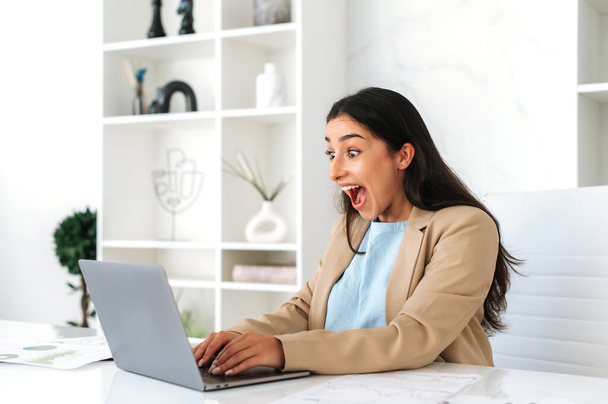 Amazed joyful happy indian or arabian business woman in elegant clothes, sitting in modern office, happily looks at laptop screen, rejoicing promotion at work, big profit, success, laughs - Photo, Image