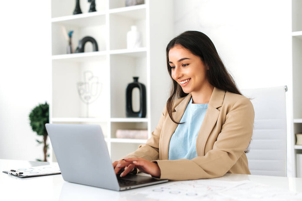 Photo of elegant successful indian or arabian business lady, company employee sits at workplace in the office, using laptop, working on a project, browsing and searching information, smiling - Photo, Image