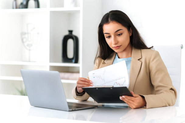 Сконцентрированная серьезная индийская или арабская деловая женщина, финансовый менеджер, сидящая на рабочем месте в офисе, использующая ноутбук, делающая бумажную работу, сосредоточенная на изучении документов, составляющая бизнес-план - Фото, изображение
