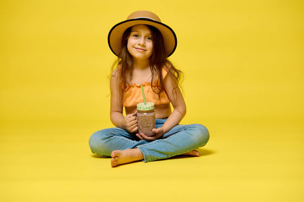 Portrait of an adorable cheerful little kid girl holding a freshly squeezed fruity juice, expressing positive emotions, smiling looking at the camera, sitting isolated over yellow studio background - Photo, Image