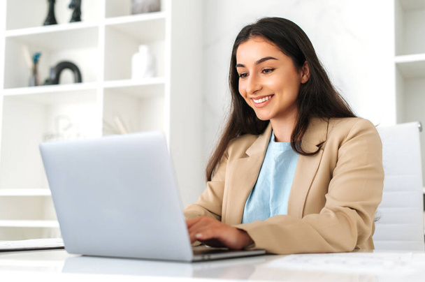 Working on a laptop. Positive successful indian or arabian business woman, company employee sitting at workplace in the office, using laptop, working on a project, looks at screen, smiling friendly - Photo, Image