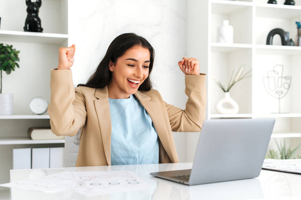 Победа, успех. Радостная изумленная счастливая индийская или арабская деловая женщина, сидящая в современном офисе, радуясь продвижению на работе, большой прибыли, радостно бросает документы, веселится, смеется, жестикулирует руками - Фото, изображение