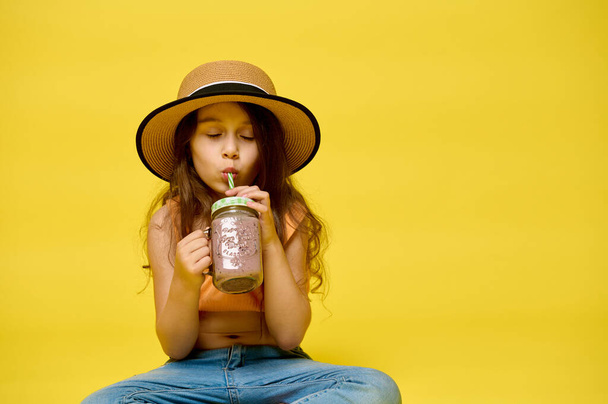 Authentic portrait of Caucasian n adorable child girl in straw summer hat, drinking cocktail from straw, enjoying her happy vacations, posing with her eyes closed on yellow isolated studio background - Photo, Image