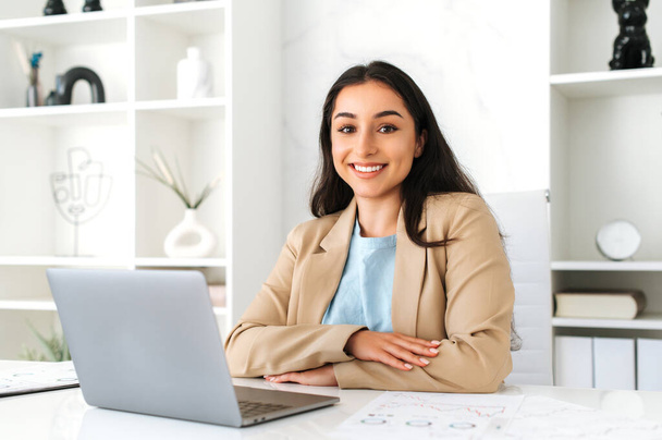 Güzel, pozitif, başarılı Hintli ya da Arap iş kadını, şirket dijital uzmanı, ofiste dizüstü bilgisayarla oturuyor, bir proje üzerinde çalışıyor, kameraya bakıyor, arkadaşça gülümsüyor. - Fotoğraf, Görsel