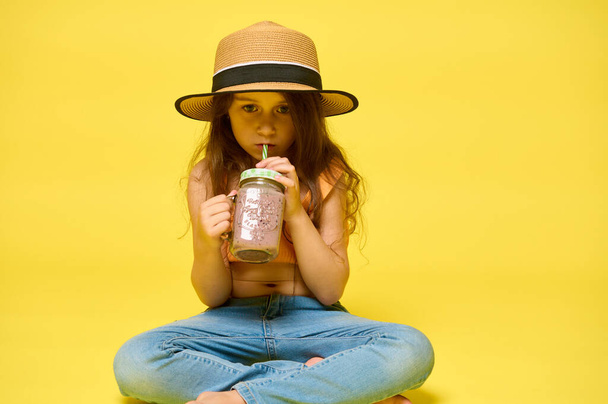 Aranyos, stílusos kislány iszik egy koktélt egy üvegpalackból szívószállal, magabiztosan néz a kamerába, elszigetelve a sárga stúdió hátterétől. hirdetési hely - Fotó, kép