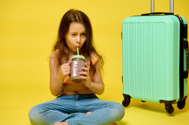 Caucasian beautiful kid girl in summer clothes, blinking while drinking lemonade or cocktail through drinking straw, sitting near her suitcase, isolated yellow studio backdrop. Beach vacation concept - Photo, Image