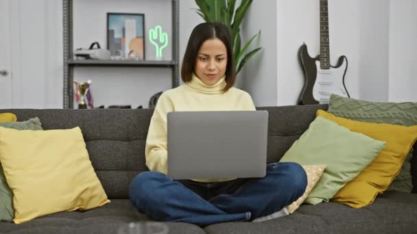 Een lachende Spaanse vrouw in een gele trui comfortabel met een laptop op een grijze bank binnen. - Video