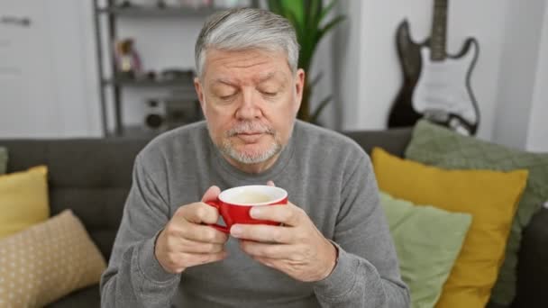 Vanhempi mies, jolla on harmaat hiukset, juo kahvia viihtyisässä olohuoneessa - Materiaali, video