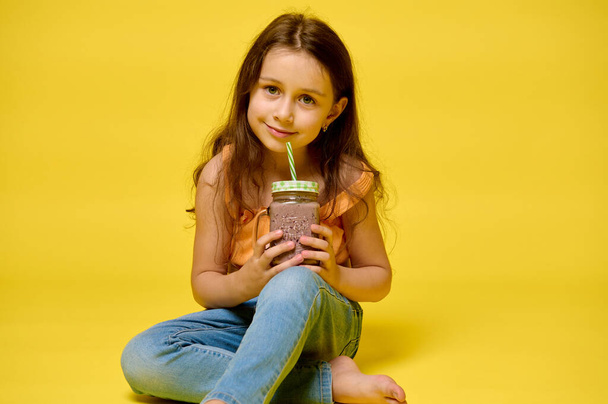 Красива дівчинка 6 років, в літньому одязі, тримає келих зі смачним коктейлем, посміхаючись дивлячись на камеру, ізольована на жовтому студійному фоні. Люди. Їжа та напої. Літня концепція - Фото, зображення