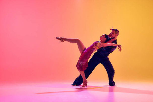 Young couple, man and woman in motion, ballroom dancers making creative performance against gradient pink yellow background in neon light. Concept of dance class, hobby, art, dance school, talent - Fotografie, Obrázek