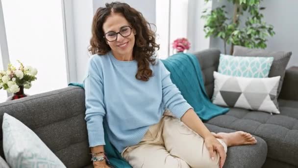Uma mulher hispânica madura conteúdo com cabelo encaracolado usando óculos senta-se relaxado em um sofá em um ambiente acolhedor sala de estar. - Filmagem, Vídeo