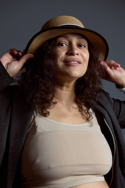 Retrato de estúdio autêntico de uma mulher grávida bonita multi étnica no topo bege, blazer cinza e um chapéu de palha, sorrindo olhando para a câmera, isolado sobre fundo de estúdio cinza moda - Foto, Imagem