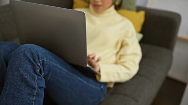 Hispanische Frau in gelbem Pullover mit Laptop, während sie drinnen auf grauem Sofa sitzt und Kopfhörer trägt. - Filmmaterial, Video