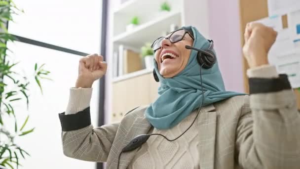 Een volwassen vrouw in een hijab viert vrolijk met opgeheven vuisten op haar werkplek met een headset op. - Video