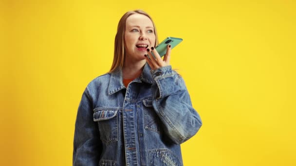 Young woman using smartphone and speaking on speakerphone against vibrant yellow studio background. Concept of beauty and fashion, self-expression, human emotions, lifestyle, connection in distance. - Footage, Video
