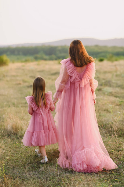 Hátsó kilátás anya és lánya ugyanabban tüll rózsaszín ruhák fogják a kezüket, és séta a természetben a nyári naplementében. Anya és lánya. Boldog gyerekkori koncepciót. Fotoshootin - Fotó, kép