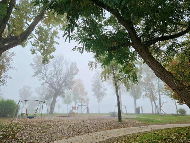 Bonita neblina e ensolarada manhã outono cenário no parque. Parque infantil para crianças em um nevoeiro. Balanços no parque infantil sob tempo nebuloso - Foto, Imagem