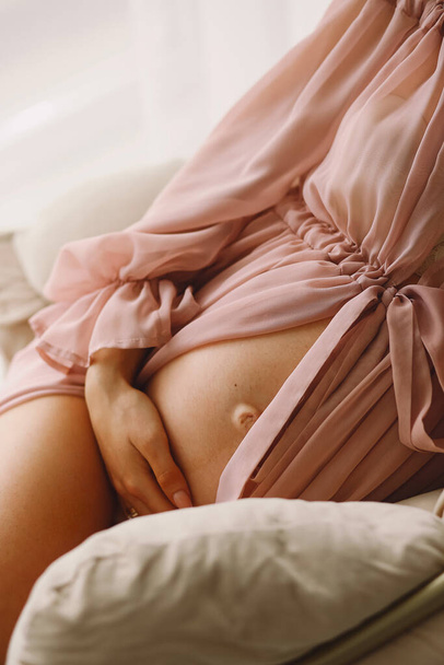 Беременная женщина в стильном розовом пеньюаре, держащая живот. Закрыть фото беременного живота. Стильная концепция материнства. Копирование текста - Фото, изображение