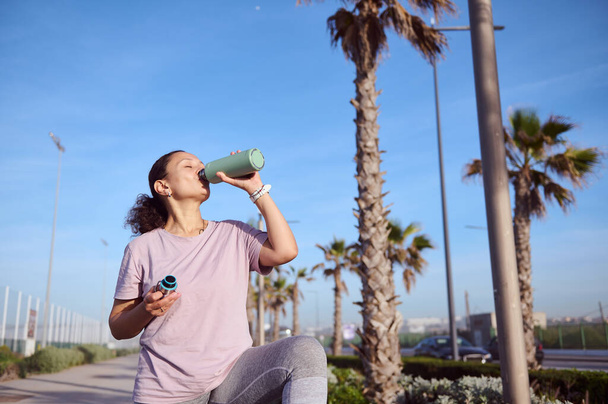 Латиноамериканская молодая спортсменка в спортивной одежде, пьет изотоническую воду, освежает после утренней пробежки по пляжу. Спорт. активный здоровый образ жизни. Похудение, похудение и диета - Фото, изображение