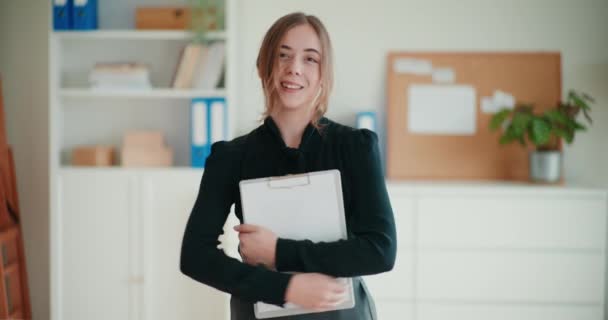 Πορτρέτο του χαμογελαστού αυτοπεποίθηση νεαρή επιχειρηματίας κρατώντας πρόχειρο ενώ στέκεται με τα χέρια σταυρωμένα σε εταιρικό χώρο εργασίας - Πλάνα, βίντεο