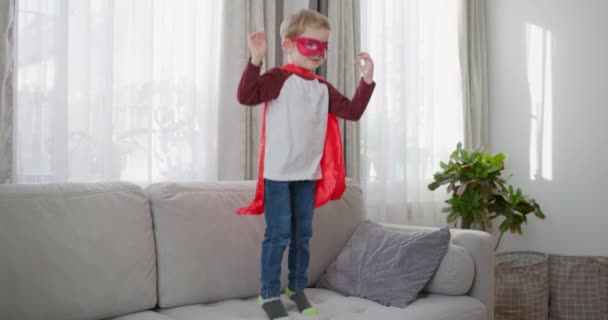 Un bambino immagina di essere un supereroe, che gioca e salta gioiosamente su un divano in un luminoso soggiorno. Filmati 4k di alta qualità - Filmati, video